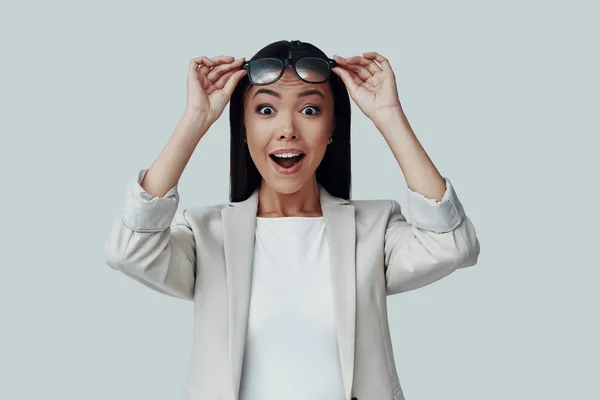 Que surpresa! Surpreendida jovem mulher asiática olhando para a câmera e sorrindo enquanto estava de pé contra fundo cinza — Fotografia de Stock