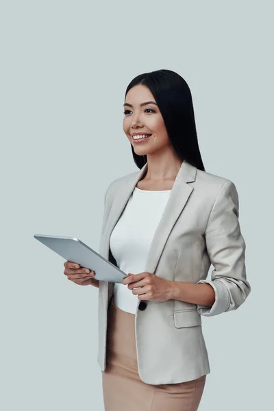 Always ready to help. Attractive young Asian woman using digital tablet and smiling while standing against grey background