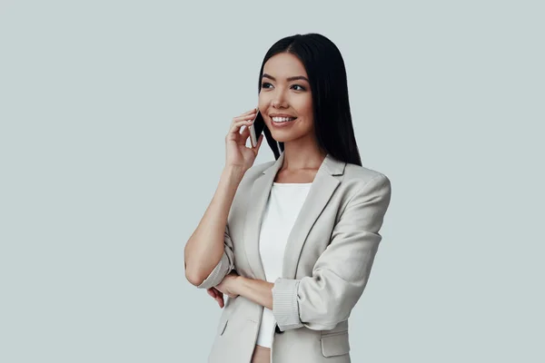 Good business talk. Attractive young Asian woman talking on the phone and smiling while standing against grey background