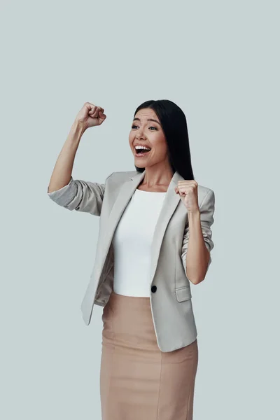 ¡Lo logré! Atractiva joven mujer asiática haciendo gestos y sonriendo mientras está de pie sobre fondo gris —  Fotos de Stock