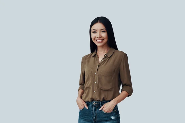Joven y hermosa. Atractiva joven mujer asiática mirando a la cámara y sonriendo mientras está de pie sobre fondo gris —  Fotos de Stock