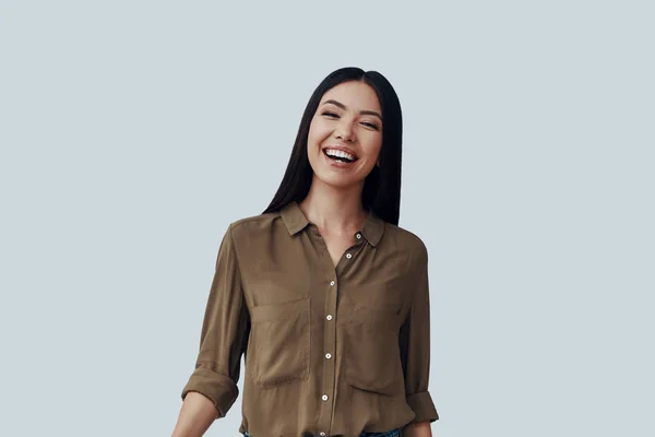 ¡Tan feliz! Hermosa mujer asiática joven mirando a la cámara y sonriendo mientras está de pie sobre fondo gris —  Fotos de Stock