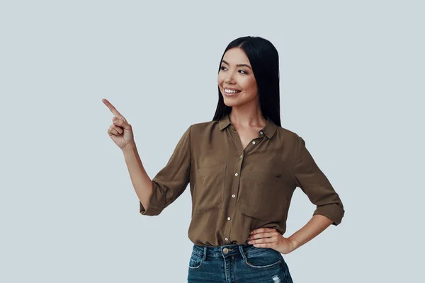 Señalando algo. Hermosa mujer asiática joven apuntando espacio de copia y sonriendo mientras está de pie sobre fondo gris —  Fotos de Stock