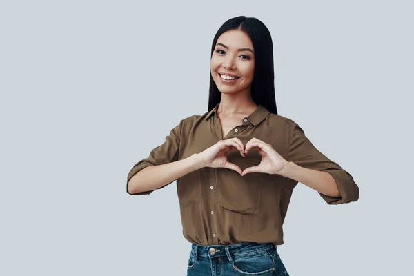 Forma de corazón. Hermosa mujer asiática joven mirando a la cámara y sonriendo mientras está de pie sobre fondo gris —  Fotos de Stock