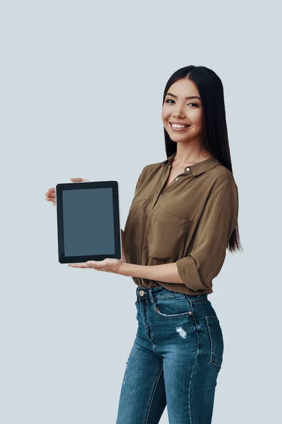 ¡Mira aquí! Hermosa mujer asiática joven mirando a la cámara y sonriendo mientras está de pie sobre fondo gris —  Fotos de Stock