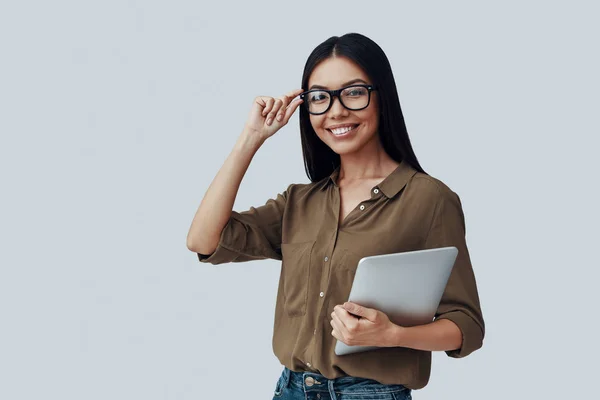 Joven e inteligente. Atractiva joven mujer asiática mirando a la cámara y sonriendo mientras está de pie sobre fondo gris —  Fotos de Stock