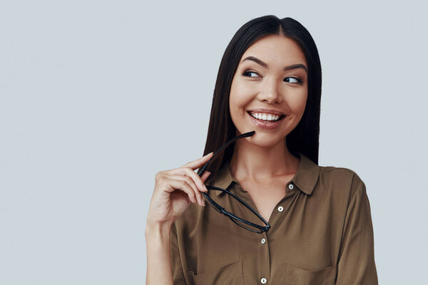 Lost in thoughts. Attractive young Asian woman looking away and smiling while standing against grey background
