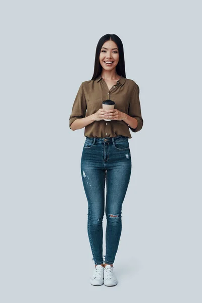 Café caliente. Longitud completa de atractiva joven mujer asiática mirando a la cámara y sonriendo mientras está de pie sobre fondo gris —  Fotos de Stock