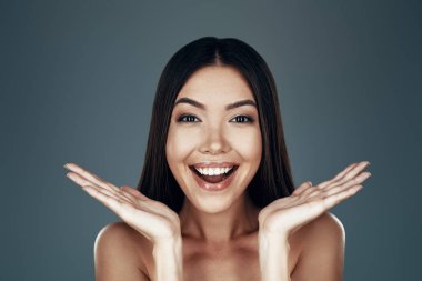 Eğlenceli. Şaşırmış genç Asyalı kadın kameraya bakıyor ve gri arka planda dururken gülümsüyor