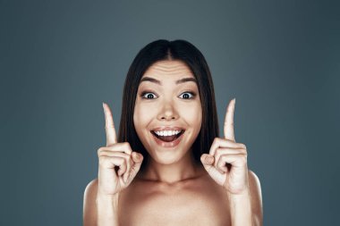 Mükemmel bir fikir. Şaşırmış genç Asyalı kadın kameraya bakıyor ve gri arka planda dururken gülümsüyor