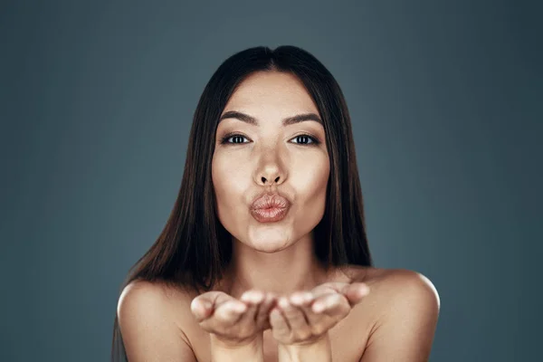 Con amor. Hermosa mujer asiática joven mirando a la cámara y soplando un beso mientras está de pie sobre fondo gris —  Fotos de Stock