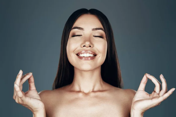 Como Zen. Hermosa mujer asiática joven manteniendo los ojos cerrados y haciendo gestos mientras está de pie sobre fondo gris —  Fotos de Stock