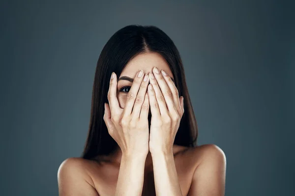 Feeling playful. Beautiful young Asian woman looking at camera and covering face with hands while standing against grey background — Stock Photo, Image