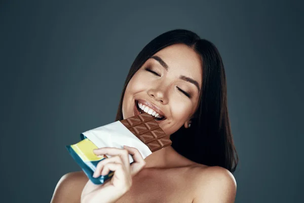 Um verdadeiro prazer. bela jovem ásia mulher sorrindo e comer chocolate enquanto em pé contra cinza fundo — Fotografia de Stock