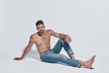 Yakışıklı genç gömleksiz adam gri arka plan karşı otururken kameraya bakıyor