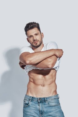 Yakışıklı genç adam kameraya bakıyor ve gri arka planda dururken soyunmak