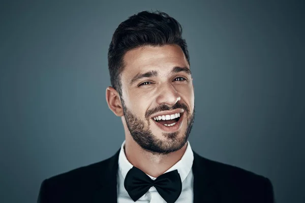 Encantador joven con corbata de lazo mirando a la cámara y sonriendo mientras está de pie sobre un fondo gris — Foto de Stock