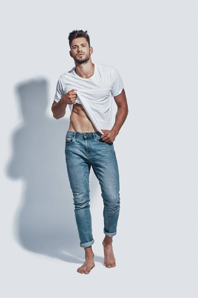 Полная длина красивого молодого человека, смотрящего в камеру и раздевающегося, стоя на сером фоне
