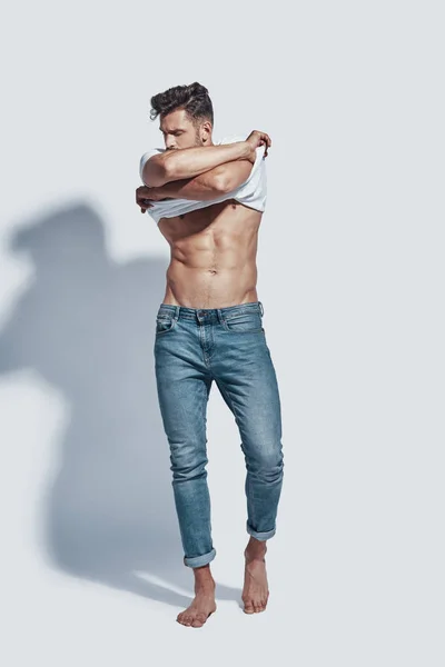 Longitud completa de joven guapo quitándose la camiseta mientras está de pie sobre fondo gris —  Fotos de Stock