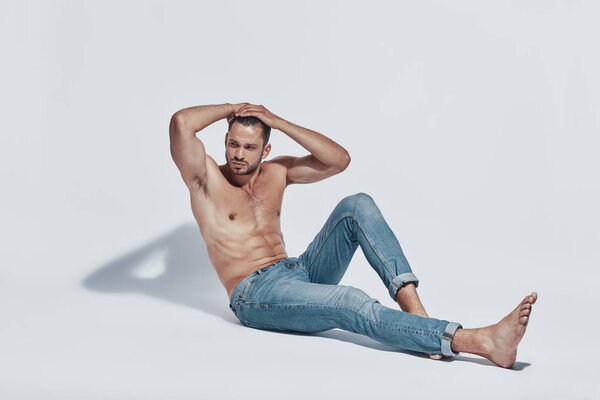 Красивый молодой мужчина без рубашки смотрит в сторону, сидя на сером фоне
