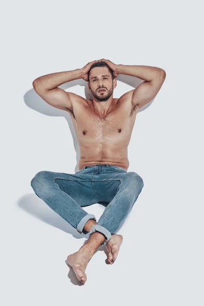 Handsome young shirtless man looking at camera and keeping hands behind hand while sitting against grey background — Stock Photo, Image