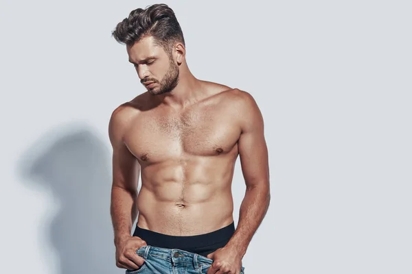 Guapo joven sin camisa hombre quitándose los pantalones vaqueros, mientras que de pie sobre fondo gris — Foto de Stock