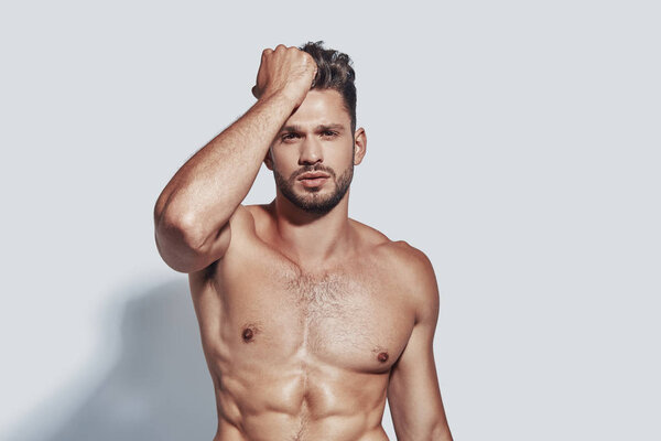 Красивый молодой мужчина без рубашки смотрит в камеру, стоя на сером фоне
