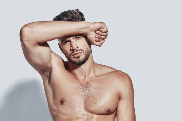 Красивый молодой мужчина без рубашки смотрит в камеру, стоя на сером фоне
