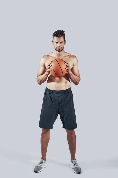 バスケットボールボールを運び、灰色の背景に立ちながらカメラを見ているスポーツ服を着たハンサムな若者の完全な長さ — ストック写真