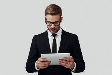 Gri arka plan aleyhine dururken dijital tablet kullanarak çalışan formalgiyim yakışıklı genç adam