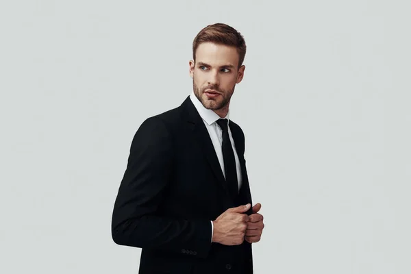 Hombre joven guapo en ropa formal mirando hacia otro lado y ajustando la chaqueta mientras está de pie sobre fondo gris — Foto de Stock