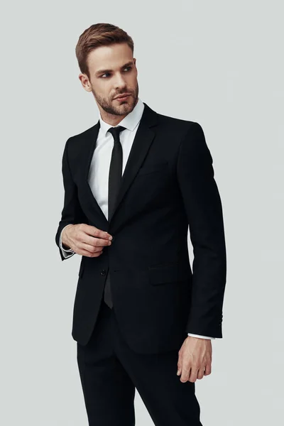 Hombre joven guapo en ropa formal mirando hacia otro lado y ajustando la chaqueta mientras está de pie sobre fondo gris — Foto de Stock