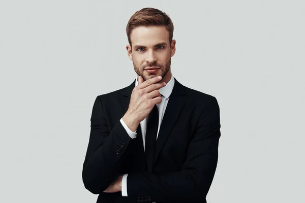 Hombre joven guapo en ropa formal mirando a la cámara y manteniendo la mano en la barbilla mientras está de pie sobre un fondo gris — Foto de Stock