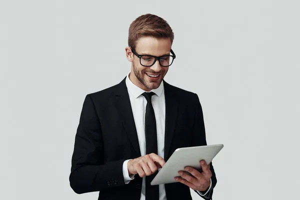 Bonito jovem em roupa formal que trabalha usando tablet digital enquanto está de pé contra fundo cinza — Fotografia de Stock