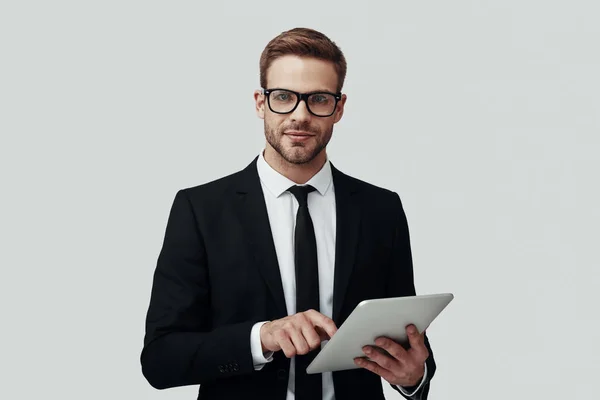 Bonito jovem em roupa formal que trabalha usando tablet digital enquanto está de pé contra fundo cinza — Fotografia de Stock