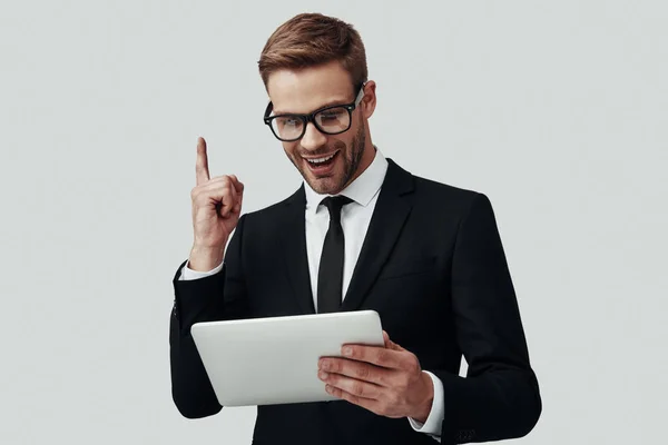 Jovem bonito em roupa formal que trabalha usando tablet digital e sorrindo enquanto está de pé contra fundo cinza — Fotografia de Stock