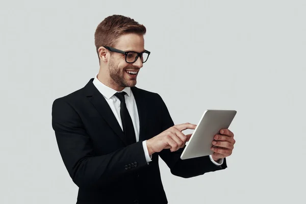 Jovem bonito em roupa formal que trabalha usando tablet digital e sorrindo enquanto está de pé contra fundo cinza — Fotografia de Stock