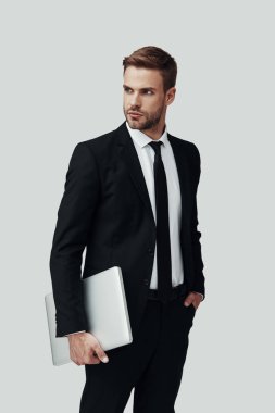 Dizüstü bilgisayar taşıyan ve gri arka plan aleyhine dururken uzağa bakan formalwear yakışıklı genç adam