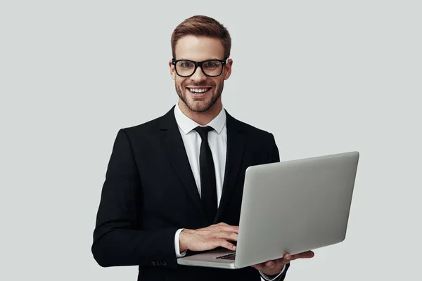 Bonito jovem em roupa formal que trabalha usando laptop e sorrindo enquanto está de pé contra fundo cinza — Fotografia de Stock