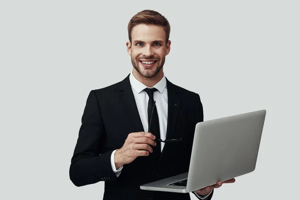 Bonito jovem em roupa formal que trabalha usando laptop e sorrindo enquanto está de pé contra fundo cinza — Fotografia de Stock