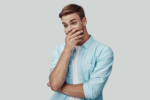 Überraschter junger Mann bedeckt Mund mit der Hand, während er vor grauem Hintergrund steht — Stockfoto