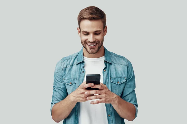 Bonito jovem usando telefone inteligente e sorrindo enquanto está de pé contra fundo cinza — Fotografia de Stock