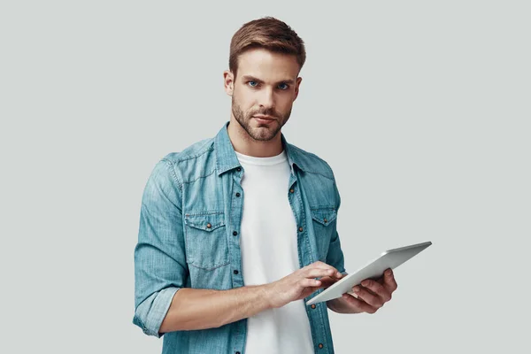 Bonito jovem olhando para a câmera e usando tablet digital enquanto está de pé contra fundo cinza — Fotografia de Stock