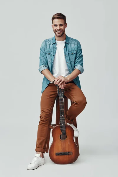 Guapo joven guitarrista sonriendo y mirando a la cámara mientras está sentado sobre un fondo gris —  Fotos de Stock