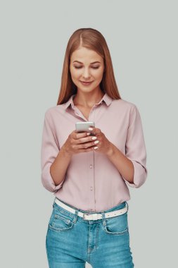 Akıllı telefon kullanan ve gri arka planda dururken gülümseyen çekici genç kadın.