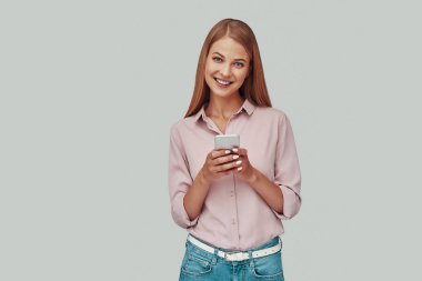 Akıllı telefon kullanan ve gri arka planda dururken gülümseyen çekici genç kadın.