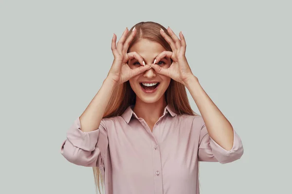 Mujer joven juguetona mirando a la cámara y sonriendo mientras está de pie sobre un fondo gris —  Fotos de Stock