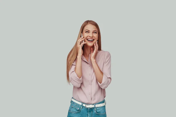 Mujer joven atractiva hablando en el teléfono inteligente y sonriendo mientras está de pie sobre fondo gris —  Fotos de Stock