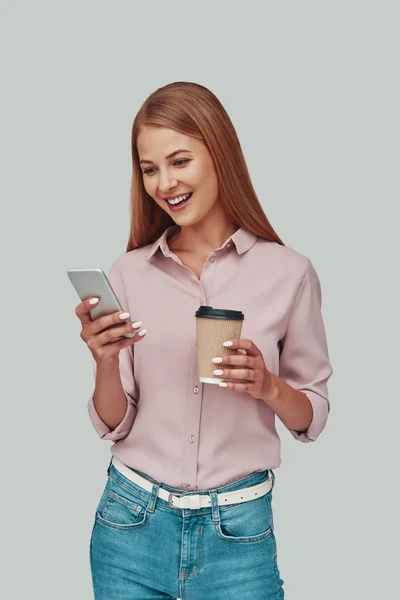 Привлекательная молодая женщина, использующая смартфон и улыбающаяся стоя на сером фоне — стоковое фото
