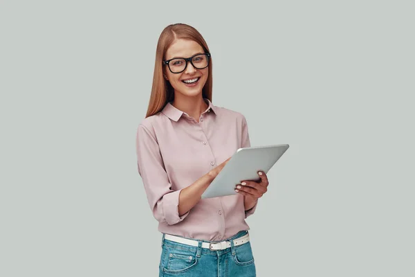 Ελκυστική νεαρή γυναίκα σε γυαλιά, χρησιμοποιώντας ψηφιακό δισκίο και χαμογελώντας ενώ στέκεται ενάντια σε γκρίζο φόντο — Φωτογραφία Αρχείου